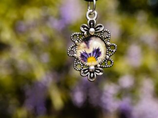 Nahaufnahme eines handgefertigten Anhängers mit echter Veilchenblüte hinter einem 12 mm Glascabochon, eingefasst in eine filigrane silberfarbene Fassung mit floralem Design (30 × 20 mm).