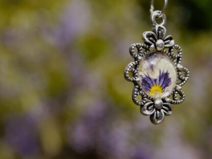 Nahaufnahme eines handgefertigten Anhängers mit echter Veilchenblüte hinter einem 12 mm Glascabochon, eingefasst in eine filigrane silberfarbene Fassung mit floralem Design (30 × 20 mm).